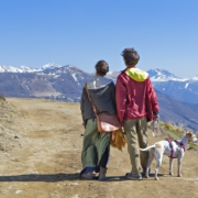 A man and woman stand, backs facing the camera, with a dog on a mountain path, looking at a view of mountains in the distance.