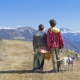 A man and woman stand, backs facing the camera, with a dog on a mountain path, looking at a view of mountains in the distance.