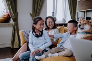 An Asian woman and two young children are smiling and playing on a yellow couch. 