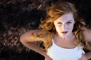 A white woman with long reddish hair laying on her back on leafy ground, looking up at the camera.