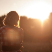 A woman looks at the sun setting behind a line of trees.