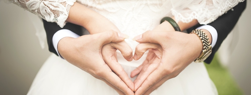 A man in a suit stands behind a woman in a wedding dress with his arms wrapped around her torso holding her hands. The couple, who we only see from the chest to the hip, is making a heart shape with their joined hands.