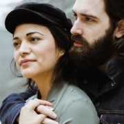 A close up of a man and woman standing together, with the woman's back to the man's chest and his arm around her front. The woman is wearing a hat and the man has a thick beard.
