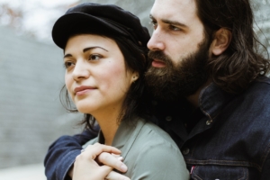 A close up of a man and woman standing together, with the woman's back to the man's chest and his arm around her front. The woman is wearing a hat and the man has a thick beard. 