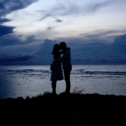 A couple standing at dusk at the edge of a body of water, leaning towards each other.