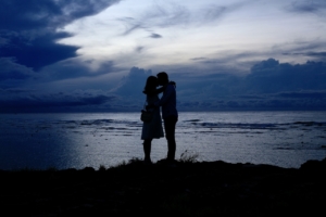 A couple standing at dusk at the edge of a body of water, leaning towards each other.