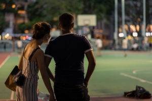 A couple standing on the side of a sports field, leaning into one another, from behind. 