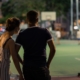 A couple standing on the side of a sports field, leaning into one another, from behind.
