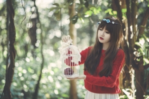 A young Asian woman stands outside in a tree filled area, holding an empty white bird cage. 