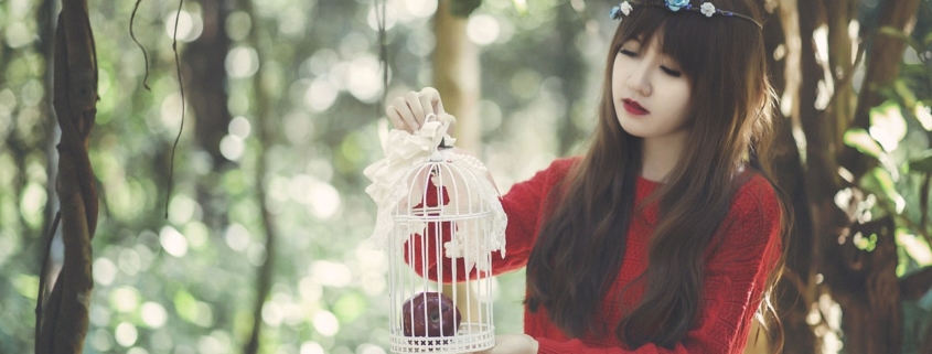 A young Asian woman stands outside in a tree filled area, holding an empty white bird cage.