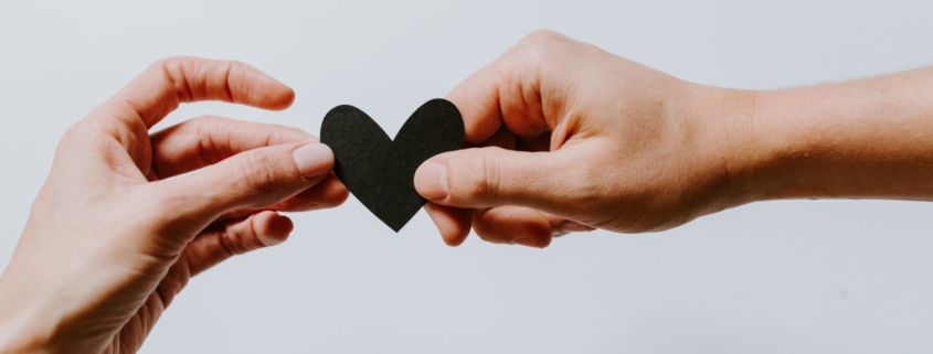 A stock photo showing two hands, reaching from the sides of the frame, holding a heart made of black paper.