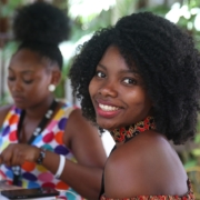 Two Black women sitting outdoors at a table with food on it. One is looking down at her plate and the other is turned toward the camera, smiling.