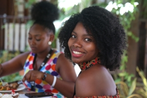 Two Black women sitting outdoors at a table with food on it. One is looking down at her plate and the other is turned toward the camera, smiling. 