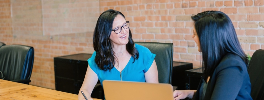 Two dark haired women talking and sitting in black chairs at a long wooden conference table that's in front of a red brick wall. There are papers and a laptop on the table.