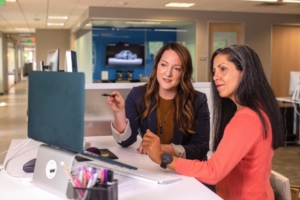 Two white women sitting at a desk, looking at a computer monitor.