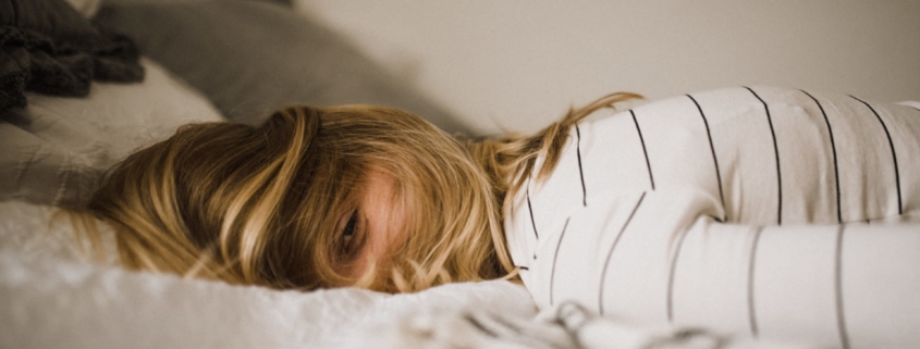A white, blonde woman laying face down on a bed with her hair covering her face.