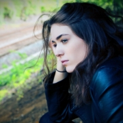 A stock photo of a young white woman with dark hair and a dark jacket on, standing outside looking thoughtful.