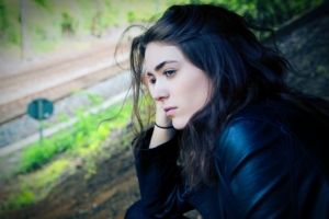 A stock photo of a young white woman with dark hair and a dark jacket on, standing outside looking thoughtful. 