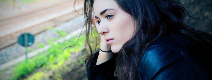 A stock photo of a young white woman with dark hair and a dark jacket on, standing outside looking thoughtful.
