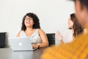 A stock photo of a Black woman sitting with colleages at a conference room table with a laptop. She is laughing.