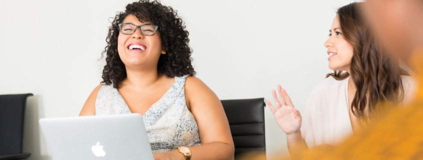 A stock photo of a Black woman sitting with colleages at a conference room table with a laptop. She is laughing.