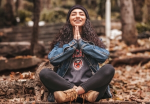 A stock photo of a young woman sitting cross legged in fallen leaves, holding her hands together like she's praying or meditating, smiling.