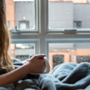 A photo of a person with long hair laying under a blanket, holding a mug, looking out a window.