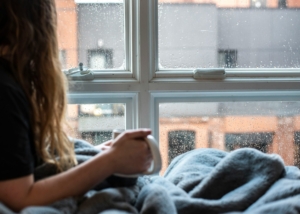 A photo of a person with long hair laying under a blanket, holding a mug, looking out a window.