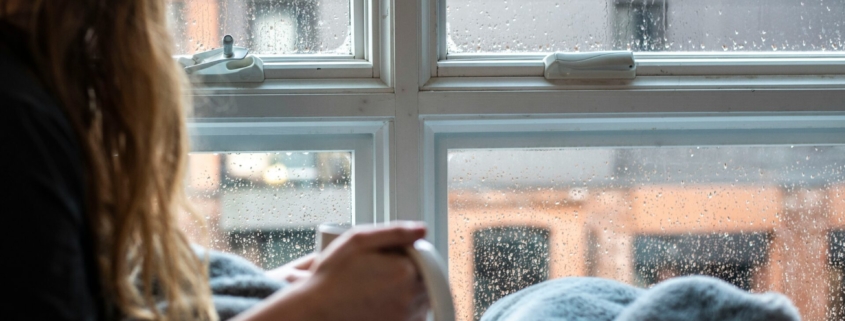 A photo of a person with long hair laying under a blanket, holding a mug, looking out a window.