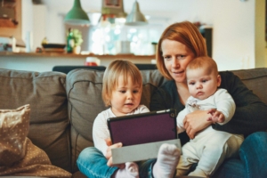 A woman with red hair sitting on a couch with two young children, looking at a tablet.