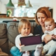 A woman with red hair sitting on a couch with two young children, looking at a tablet.