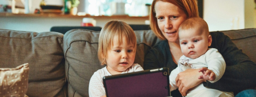 A woman with red hair sitting on a couch with two young children, looking at a tablet.