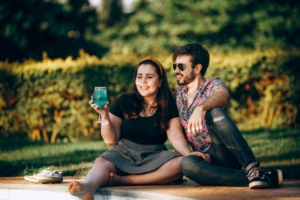 A man and a woman sitting down next to each other on grass, talking. The woman has a beverage in her hand.