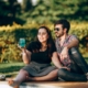 A man and a woman sitting down next to each other on grass, talking. The woman has a beverage in her hand.