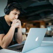 A man wearing headphones, looking at his computer with his hands clasped in front of his mouth.