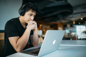 A man wearing headphones, looking at his computer with his hands clasped in front of his mouth.