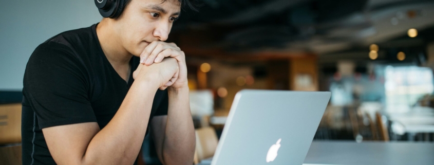 A man wearing headphones, looking at his computer with his hands clasped in front of his mouth.