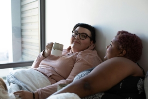 Two plus size women drinking coffee in bed together.