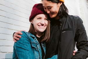 Two women wearing jackets and beanies embracing outside a building. 