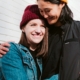 Two women wearing jackets and beanies embracing outside a building.