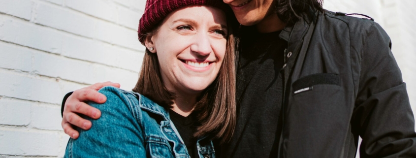 Two women wearing jackets and beanies embracing outside a building.