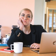 A bored or stressed looking white girl sitting a at a table in front of a laptop and coffee mug.