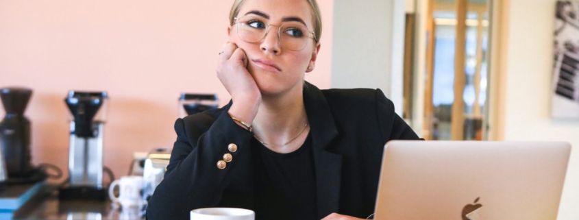 A bored or stressed looking white girl sitting a at a table in front of a laptop and coffee mug.