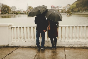 A couple from behind, standing by a body of water, each holding umbrellas.