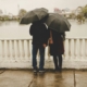 A couple from behind, standing by a body of water, each holding umbrellas.