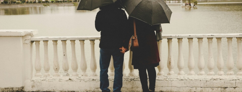 A couple from behind, standing by a body of water, each holding umbrellas.