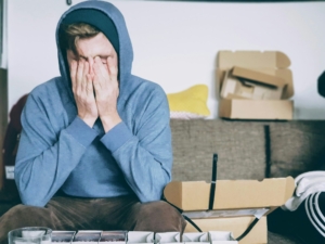 A young white man sitting on a couch surrounded by boxes, holding his face in his hands.