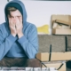 A young white man sitting on a couch surrounded by boxes, holding his face in his hands.