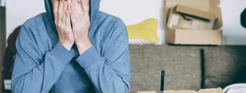 A young white man sitting on a couch surrounded by boxes, holding his face in his hands.