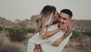 A heterosexual couple in a desert landscape, with the woman riding on the man's back. They're looking at each other affectionately.